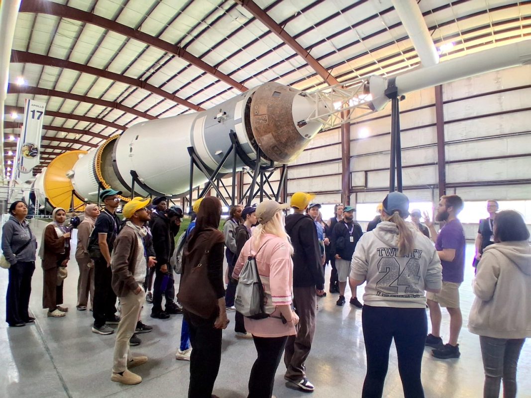 The group of 30 Level 2 and 3 Bradford College Science and Computing students stand in front of a huge model rocket on its side, while inside an aircraft hangar. Someone from NASA is speaking to the group.