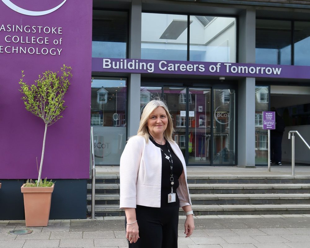 A woman stands in front of a college entrance, smiling. She is proud of her new role.