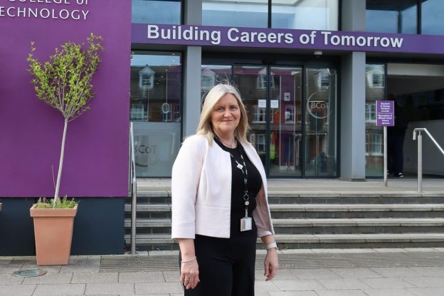 A woman stands in front of a college entrance, smiling. She is proud of her new role.