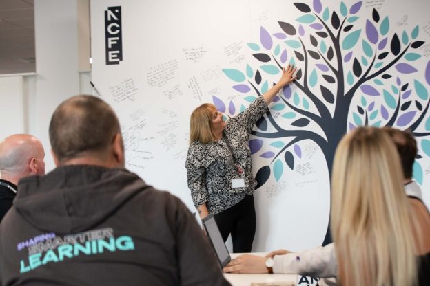 NCFE colleagues sit and watch as another colleague points to a board with an illustration of a tree