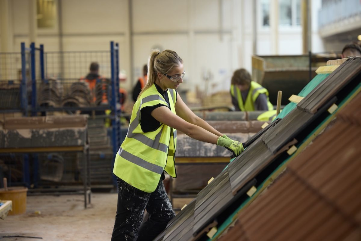 A female student in a high-vis vest and gloves places roofing tiles on a mock roof structure in a training workshop. Other students work in the background.