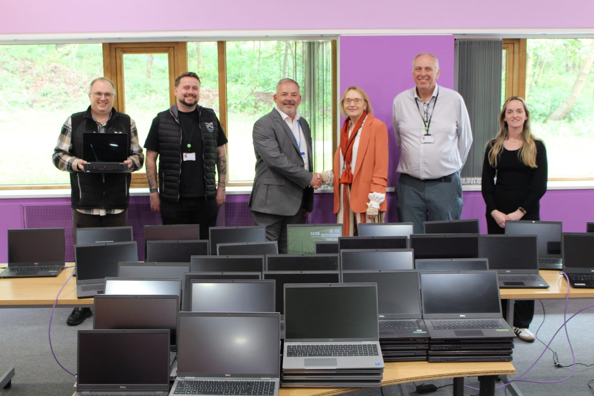 four men and two women smiling, in front of 100 laptops. a man and a woman are shaking hands.