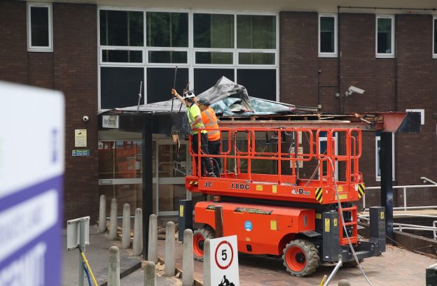 An orange cherry-picker lifts two construction workers to the top of a building's ground-floor entrance. The staff are wearing high-vis jackets and demolishing the old canopy over the entrance ready for refurbishment.