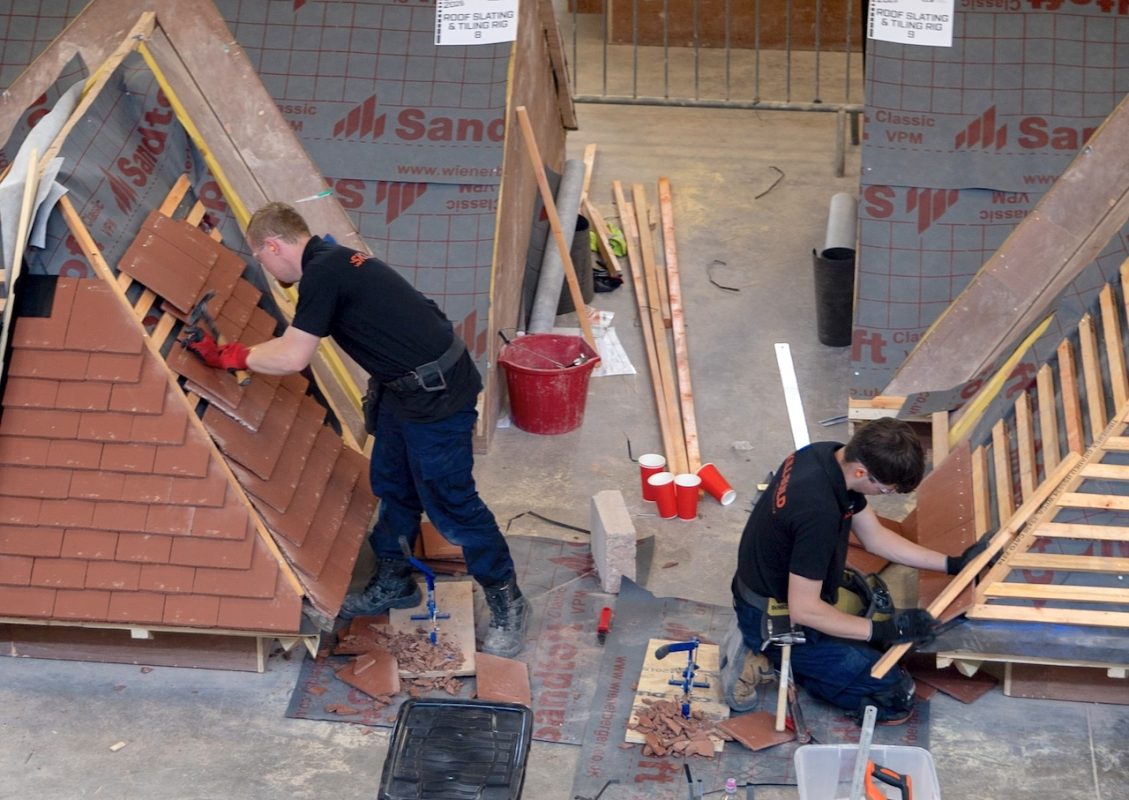 LCB competitors take part in a regional heat of SkillBuild 2026. Two male apprentices work on mock roofing rigs in a training warehouse. Both are fitting roof tiles with materials and tools around them on the floor.