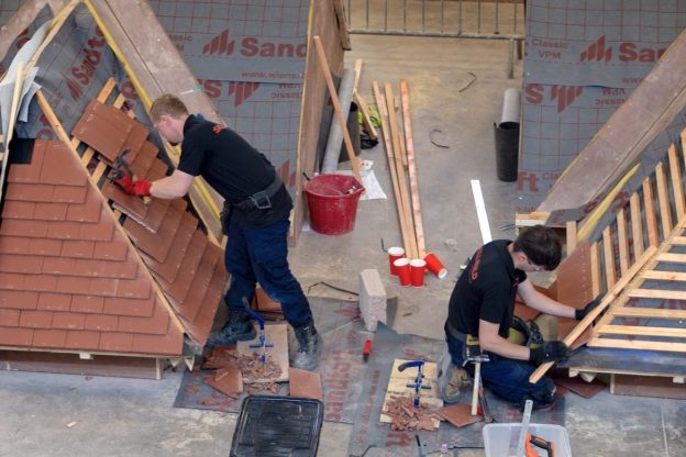 LCB competitors take part in a regional heat of SkillBuild 2026. Two male apprentices work on mock roofing rigs in a training warehouse. Both are fitting roof tiles with materials and tools around them on the floor.