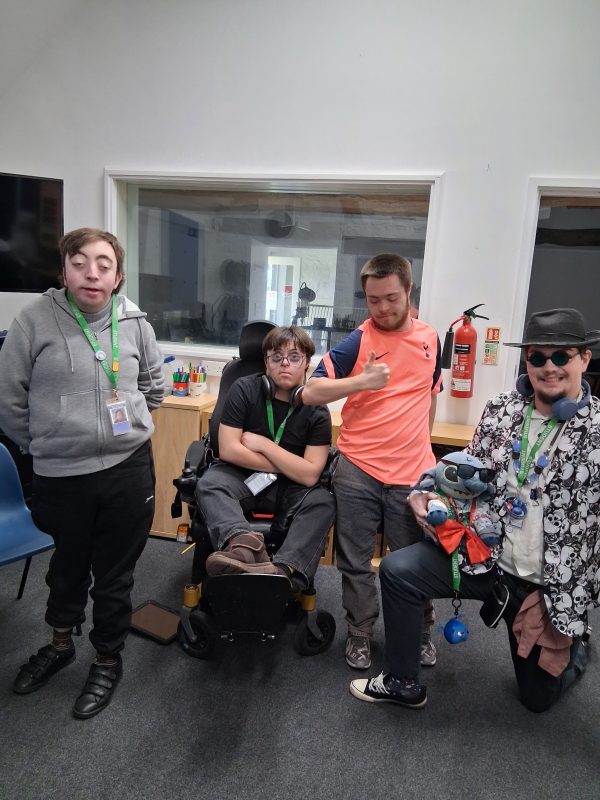 Four students from the Orpheus Centre pose for a photograph. From left is a boy in a grey hoodie, a boy in a wheelchair, a boy with a pink top and a boy kneeling with sunglasses, a hat and puppet.