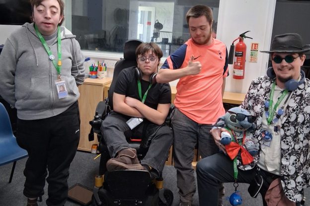 Four students from the Orpheus Centre pose for a photograph. From left is a boy in a grey hoodie, a boy in a wheelchair, a boy with a pink top and a boy kneeling with sunglasses, a hat and puppet.