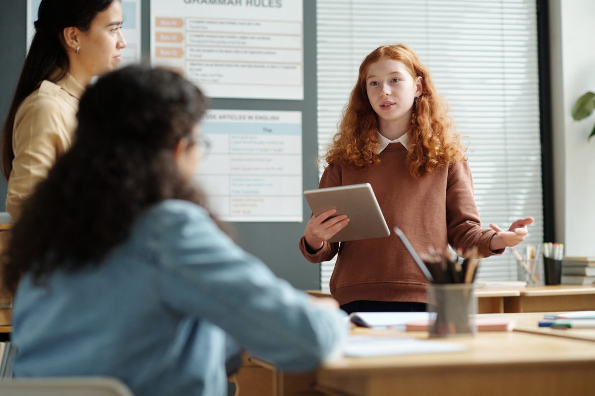 Cute confident schoolgirl with tablet standing in front of her classmates and teacher while looking at one of them and explaining new rules