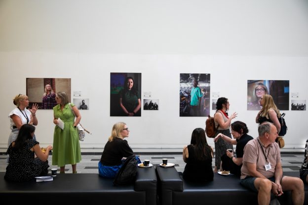 People sat on benches in front of a wall featuring a series of portraits