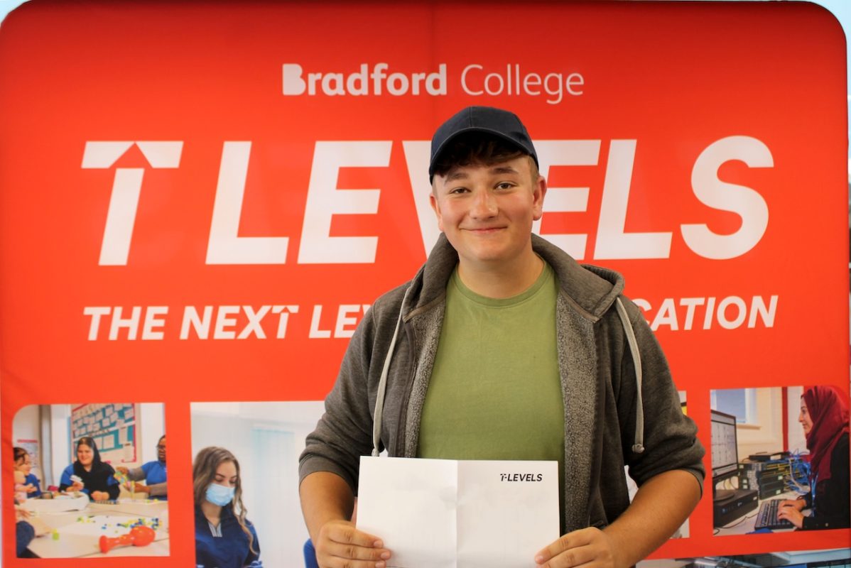 A male Bradford College T Level student collects his results. He stands in front of a red T Level banner smiling, wearing a cap and hoodie, while holding his paper certificate.