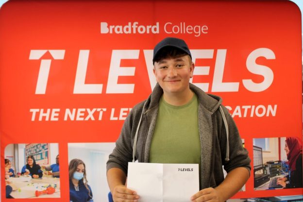 A male Bradford College T Level student collects his results. He stands in front of a red T Level banner smiling, wearing a cap and hoodie, while holding his paper certificate.