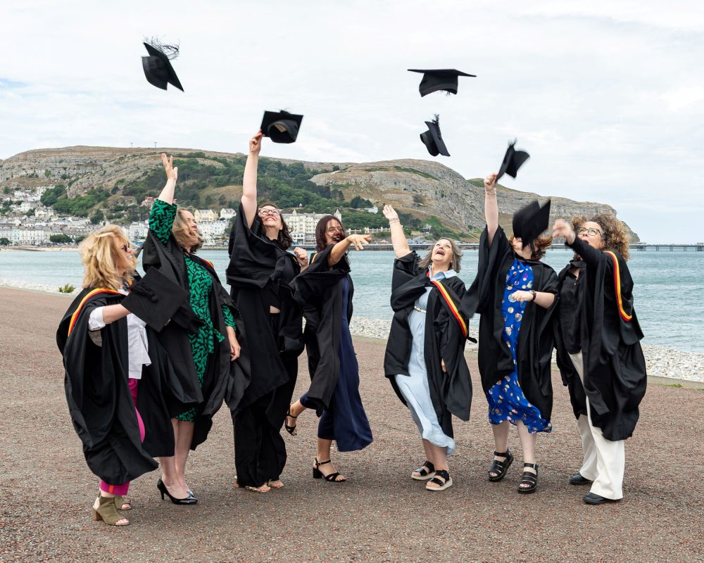 A group of students in graduation gowns and caps are standing together on the beach at Llandudno. They are throwing their caps into the air.