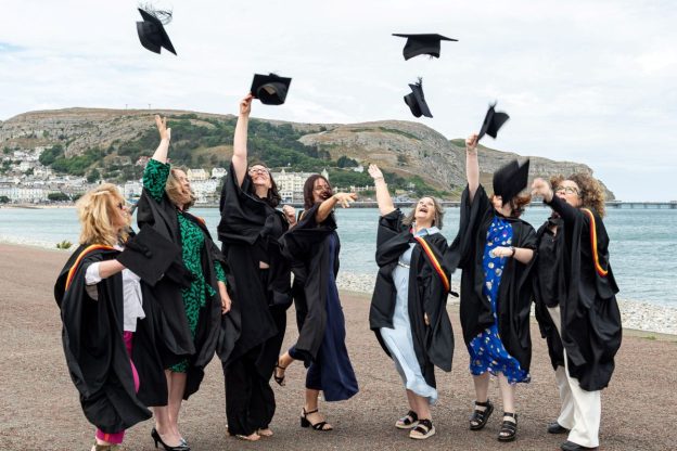 A group of students in graduation gowns and caps are standing together on the beach at Llandudno. They are throwing their caps into the air.