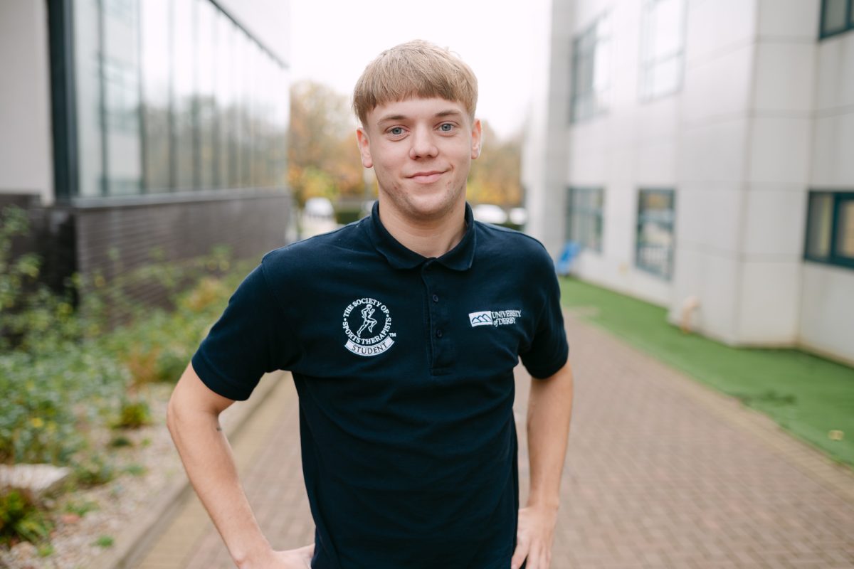 A university of derby student stands and smiles at the camera