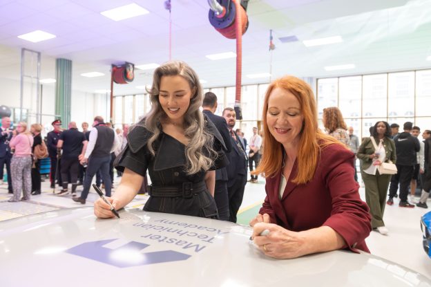 Two women sign the bonnet of a car with black pens for the photo-op. They are standing in a bright workshop with guests seen networking in the background.