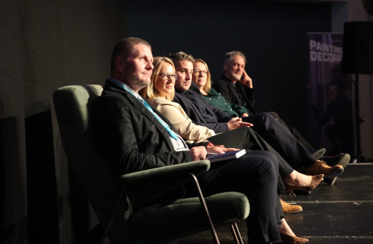 A group of male and female panelists sit in a row of arm chairs on stage. They are photographed from the side and the stage is dark but with a spotlight on the speakers.