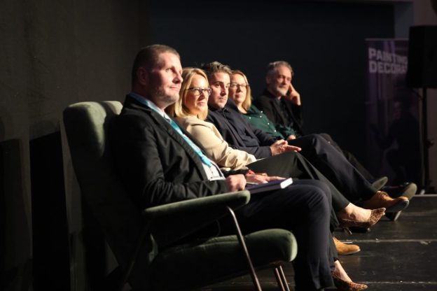 A group of male and female panelists sit in a row of arm chairs on stage. They are photographed from the side and the stage is dark but with a spotlight on the speakers.