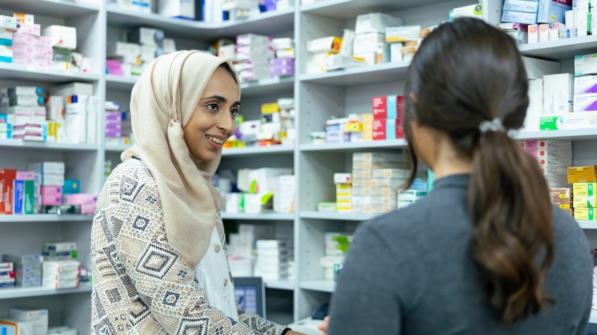 Two women stand in the back of pharmacy. One has her back to the camera and the other faces sideways wearing a headscarf. Around them are shelves of medicines.