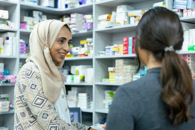 Two women stand in the back of pharmacy. One has her back to the camera and the other faces sideways wearing a headscarf. Around them are shelves of medicines.