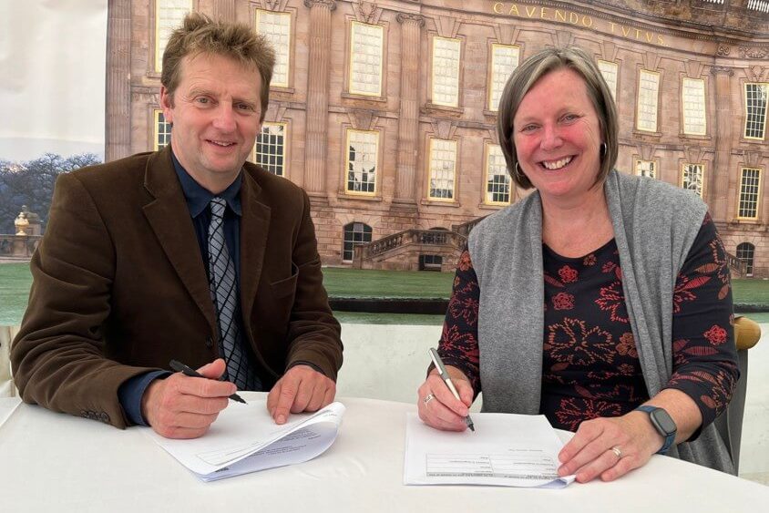 Two people are pictured in front of an image of Chatsworth House in Derbyshire